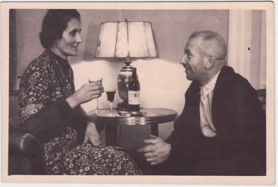 Das Schwarzweißfoto zeigt Hedwig und Emil Behr, die sich lächelnd auf Sesseln gegenüber sitzen und Rotwein trinken. Im Hintergrund steht ein Wohnzimmertischen mit einer Lampe.